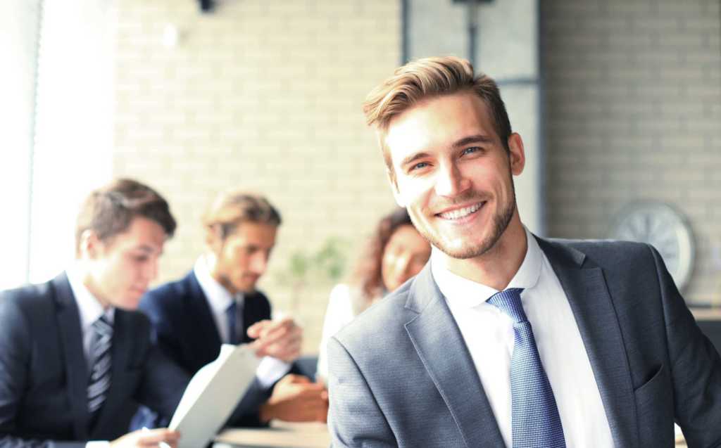 Image of young white businessman wearing a suit with a well groomed beard and hair in an office meeting smiling at the camera while three colleagues discuss business behind him.